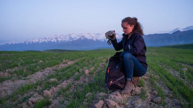 A woman is knelt on a field with a raised camera.