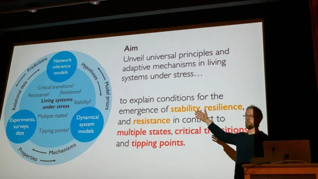 Martin Rosvall stands to the side of a large screen on which a slide is shown with images and text describing stress response modeling at IceLab