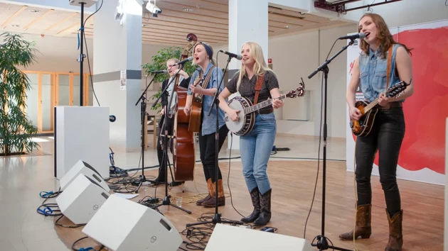 Four female artist play instruments and sing into microphones on a stage.