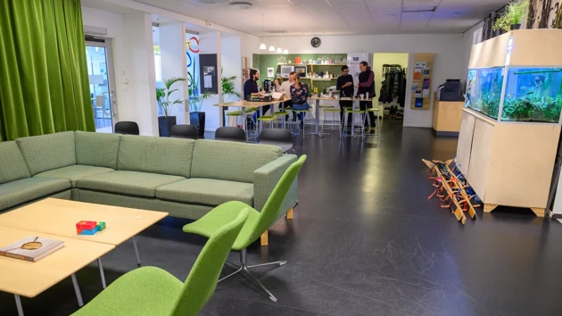 A living room and kitchen area  filled with furniture and a fish tank. A few people are gathered at the far end tables in the kitchen area.