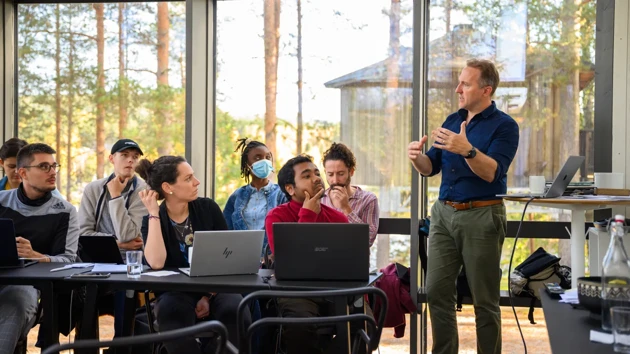 A group of students sitting at tables with laptops open listen to a man standing and speaking while gesturing with his arms. They are in a glass enclosed room, trees and another wooden house are visible outside. 