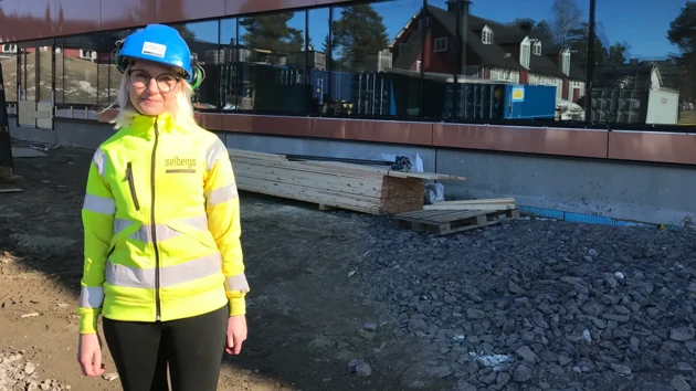 A woman with high-visibility clothing and a blue helmet looks into the camera on a building site.