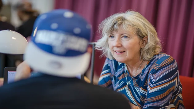 A woman is sat opposite a person in a cap and provides guidance.  