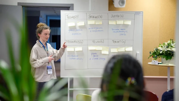 Josephine stands next to a whilte board with post-its and topics displayed in boxes.