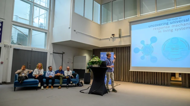 Five people sit on a blue sofa and one stands speaking next to a large screen.