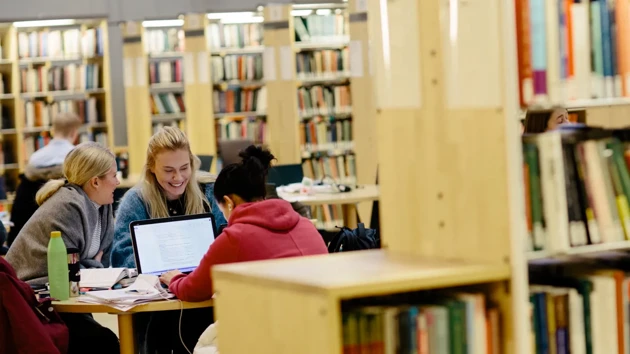 Tre personer som sitter vid ett bord i biblioteket och pluggar. Runt bordet syns flera bokhyllor med böcker.