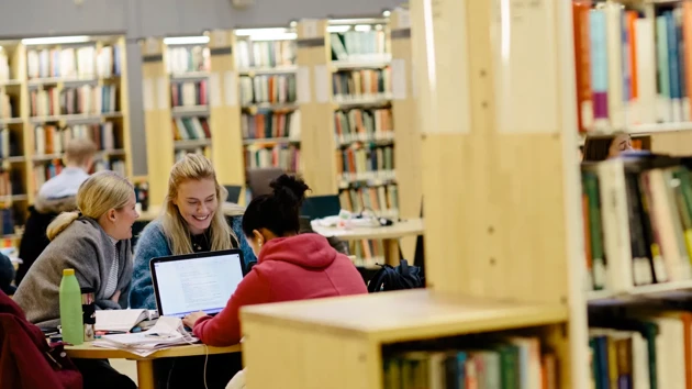 Three people sitting at a table in the library, studying. Around the table are several bookcases with books.