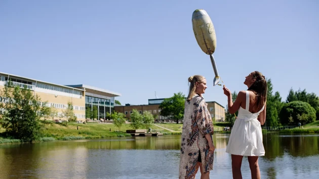 Två finklädda kvinnor står vid en damm på campus med en ballong i luften.