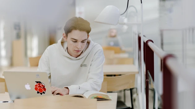 A male student is focusing on a book whilst taking notes on his laptop.
