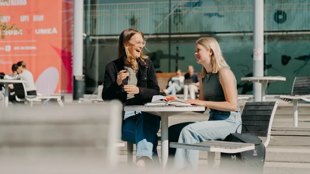 Två leende studenter som sitter vid ett utomhusbord på campus.