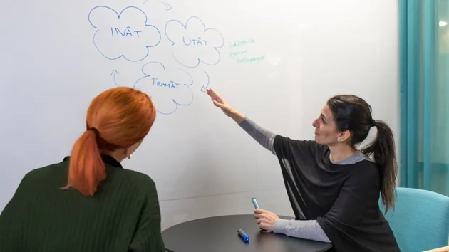 A woman sits at a small table pointing at a whiteboard with another person listening.